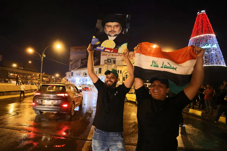 Deux hommes brandissent un drapeau irakien et un portrait de leader politique au bord d'une route en souriant.