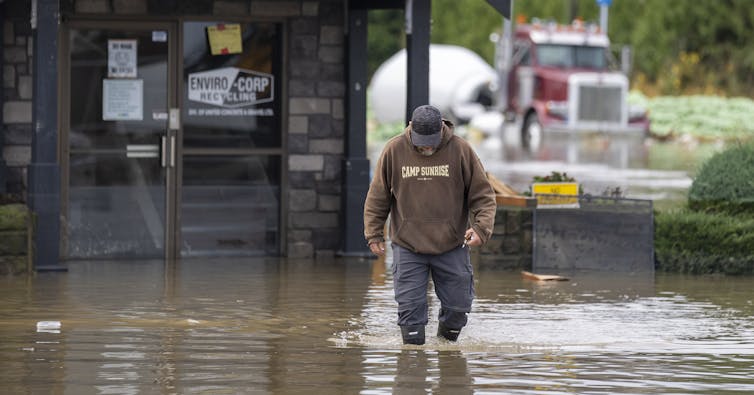 A man walking through knee-high water. In the background, a large truck and the front of a building are submerged in the water.