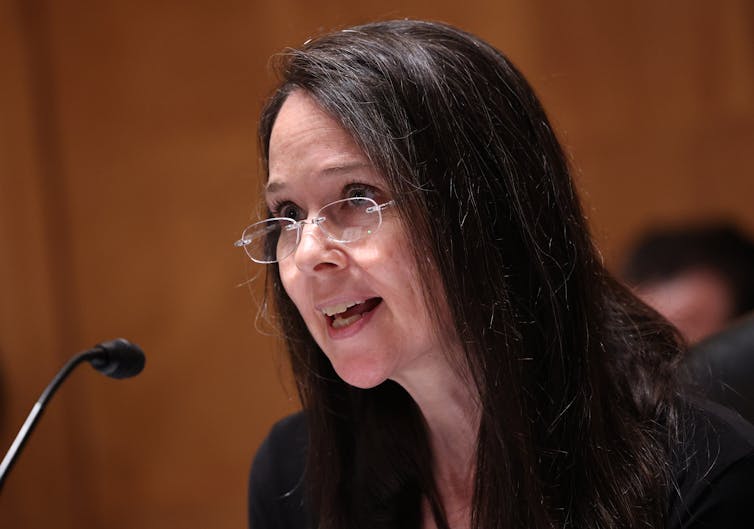 a woman with long dark hair wearing eyeglasses speaks into a microphone