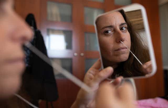 A woman holds a mirror as she conducts a nasal swab.