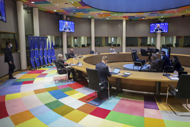 Delegates in a room with a floor made up of different coloured squares, seated several feet apart at a round table watching video screen