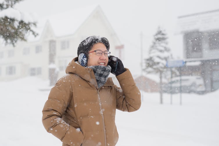 A young man on the phone outdoors in a snowstorm and snowflake earmuffs smiling