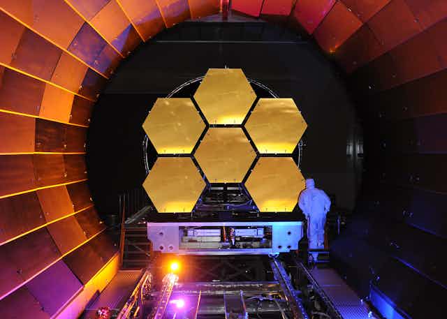 6 panneaux hexagonaux dorés reliés les un aux autres, dans un tube circulaire. Un technicien en combinaison blanche examine l'un des panneaux.
