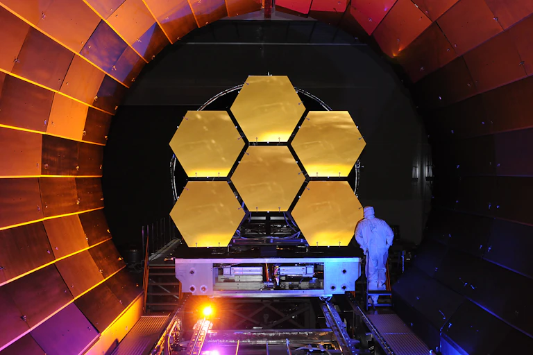 6 panneaux hexagonaux dorés reliés les un aux autres, dans un tube circulaire. Un technicien en combinaison blanche examine l'un des panneaux.