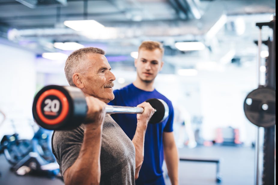 An older man lifts a barbell while his trainer looks on.