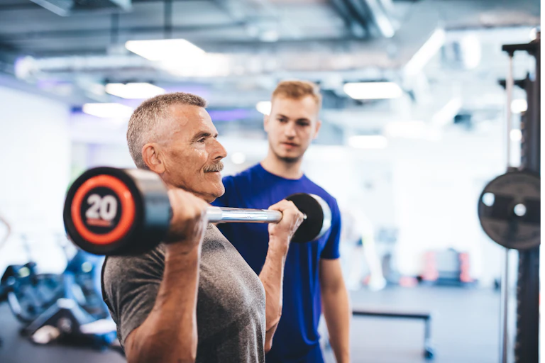 An older man lifts a barbell while his trainer looks on.