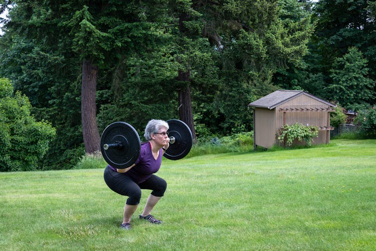 An older woman performing a barbell squat outdoors.