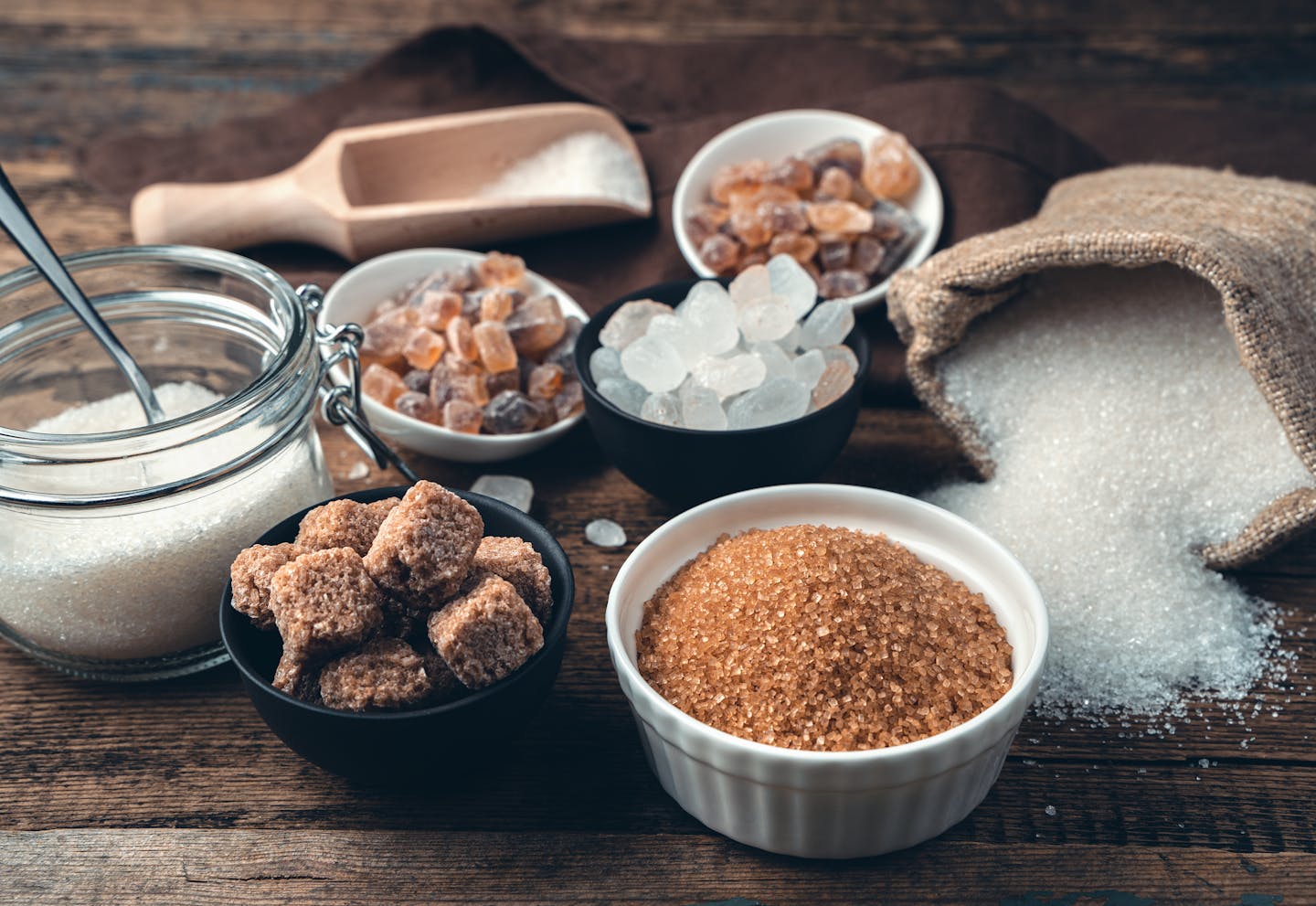 Different types of sugars in bowls, scoops and bags on a wooden table.