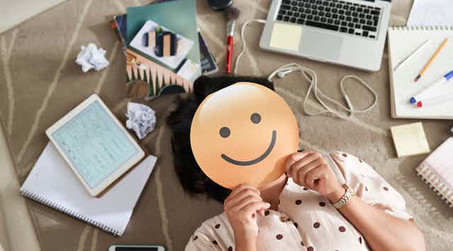 A person holds a smile emoji over their face near a laptop.