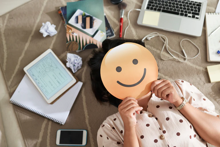 A person holds a smile emoji over their face near a laptop.