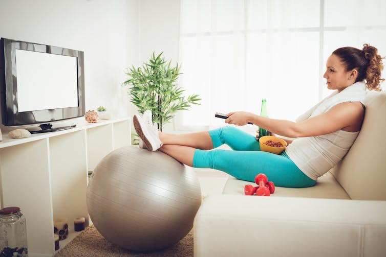 Une femme en habit d’exercice assise sur un sofa en train de regarder la télé, entourée d’équipements sportifs