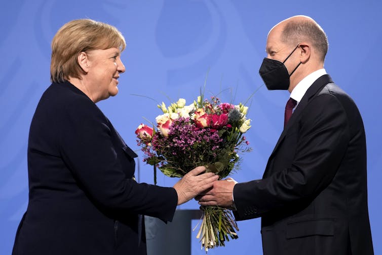 A man wearing a medical face mask handing flowers to a woman, both in front of a podium