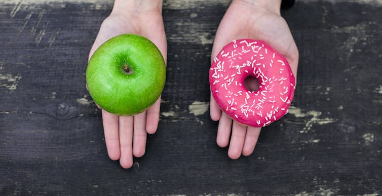 Deux mains, l’une tenant une pomme verte et l’autre un beignet avec un glaçage rose et des éclats de verre