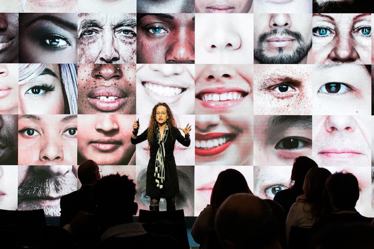 A woman stands on stage in front of an audience gesturing with her hands as the screen behind her displays a mosaic of close-up images of parts of people's faces