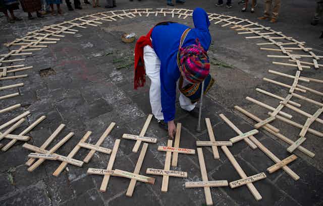 Elderly woman bending down to place wooden cross on the ground amid a circle of other wooden crosses. The crosses have names written on them.
