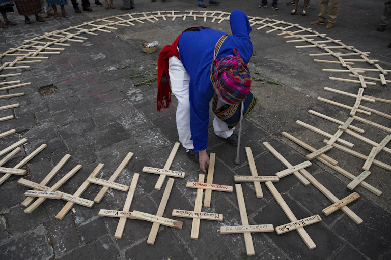 Elderly woman bending down to place wooden cross on the ground amid a circle of other wooden crosses. The crosses have names written on them.