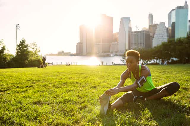 A runner stretches on grass in a park with a river behind her and a city across the water.