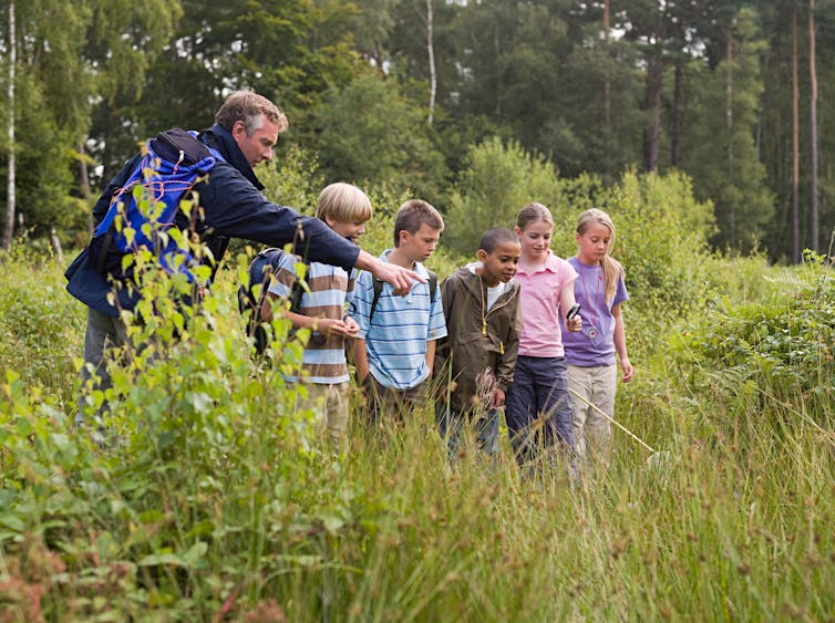 A man points to a pond where 5 children are watching with excited expressions on their faces.