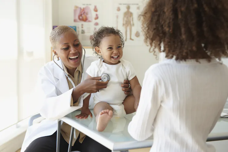 Female doctor listening to baby's breathing with stethoscope while mother looks on; all are Black.