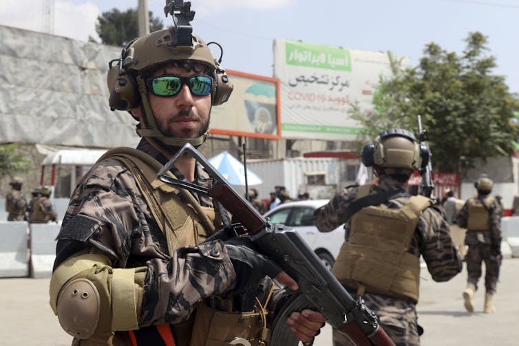 A soldier holding a gun stands in front of a troop of soldiers walking towards a building