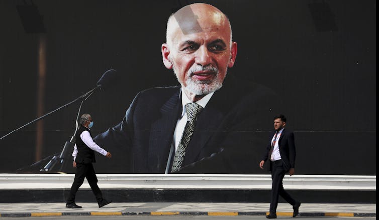 Two people walking in front of a wall with a large mural of a man's face on it.