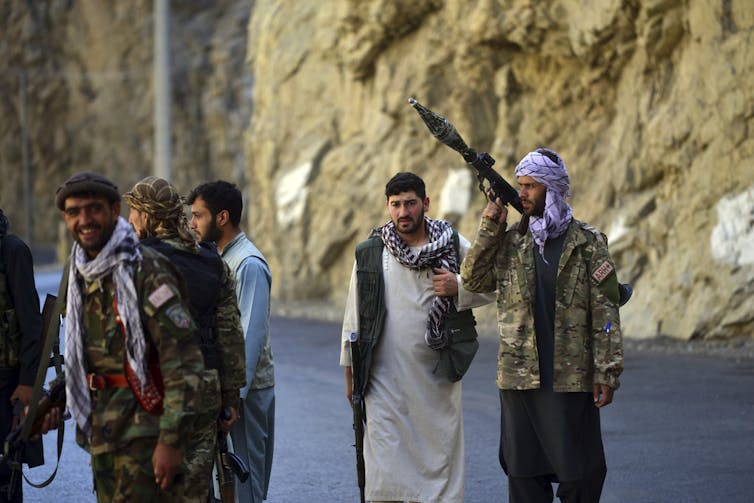 Groups men walking down a road. A few of them carry guns.