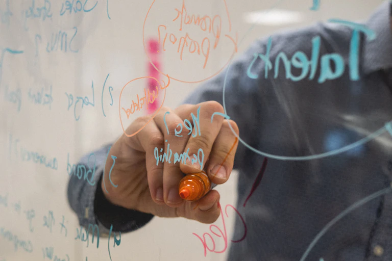 A hand holding a marker writes on a transparent whiteboard.