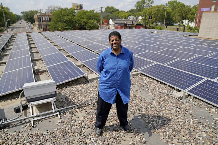 A man stands on a roof with solar panels and a community in the background.