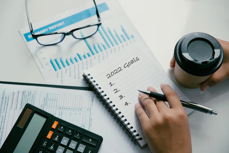 A person jots down '2022 Goals' in a notebook. In the background is a coffee cup, calculator, glasses and graph