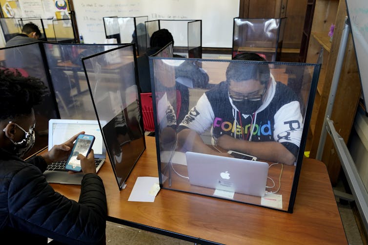 Students sit at computers, separated by clear plastic barriers