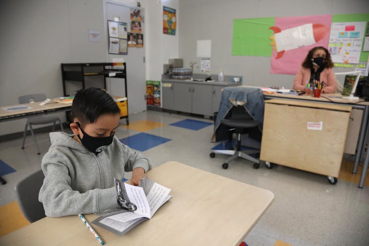 A student works at a desk while a teacher sits in the background