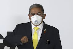 A man in a dark suit with a white shirt and yellow tie wearing a mask