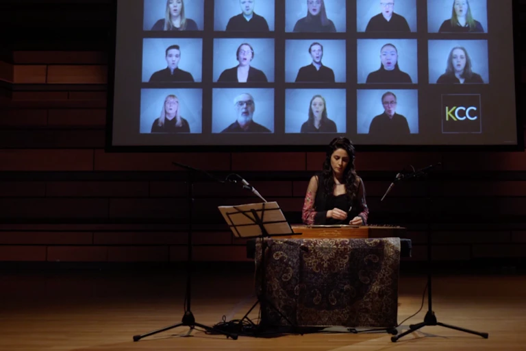 A woman is seen playing a Santur in front of a video screen showing rows of singers.