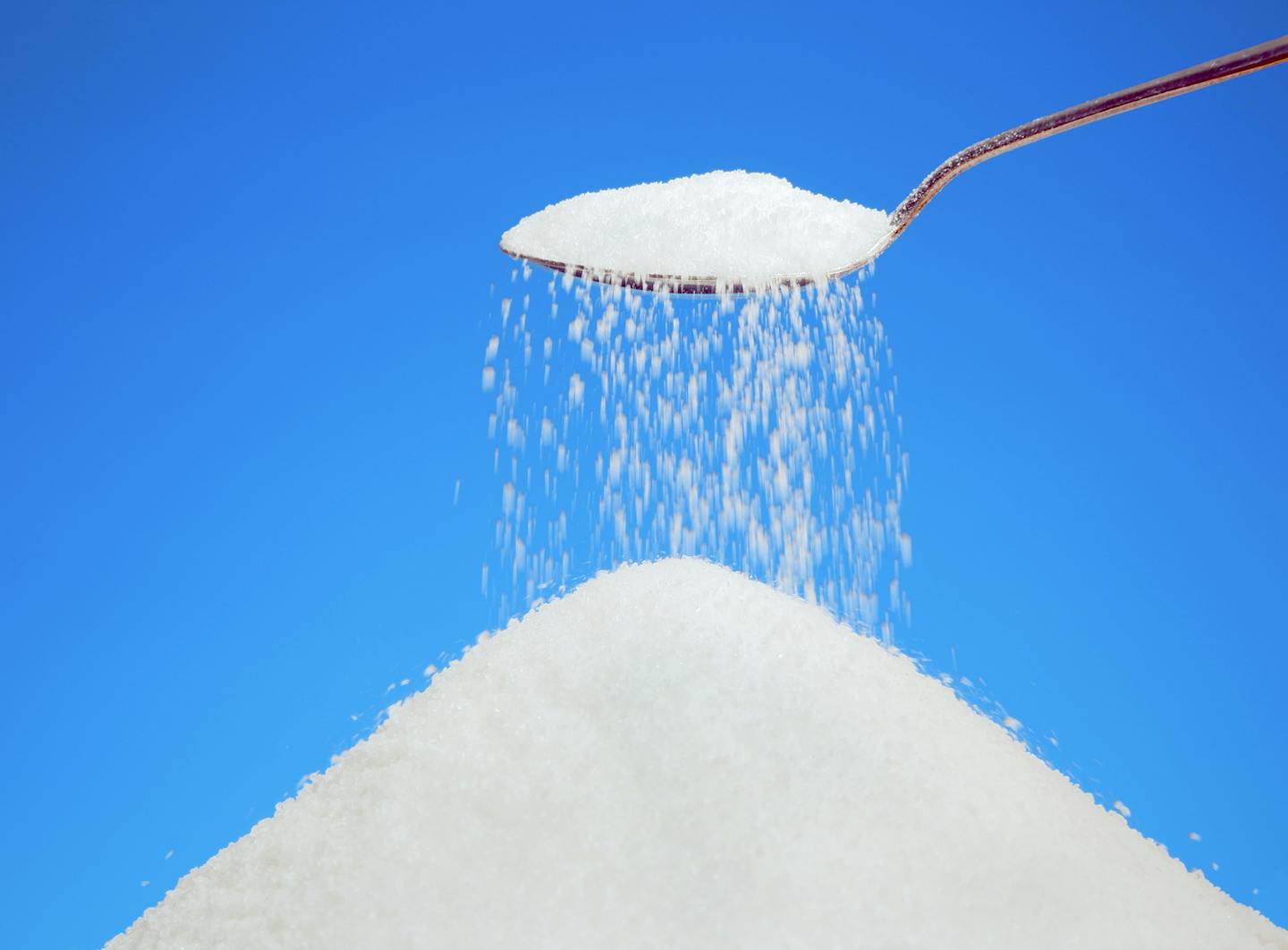 A teaspoon overflowing with sugar, held above a heap of more sugar