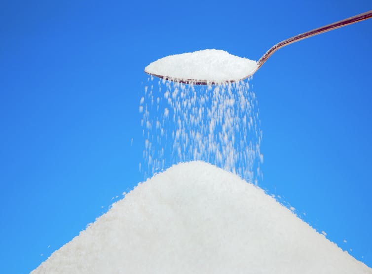 A teaspoon overflowing with sugar, held above a heap of more sugar