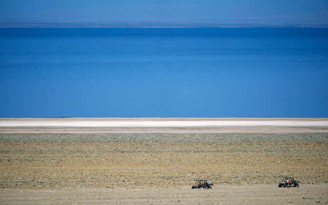 ATVs are driven along the shore of a blue body of water.