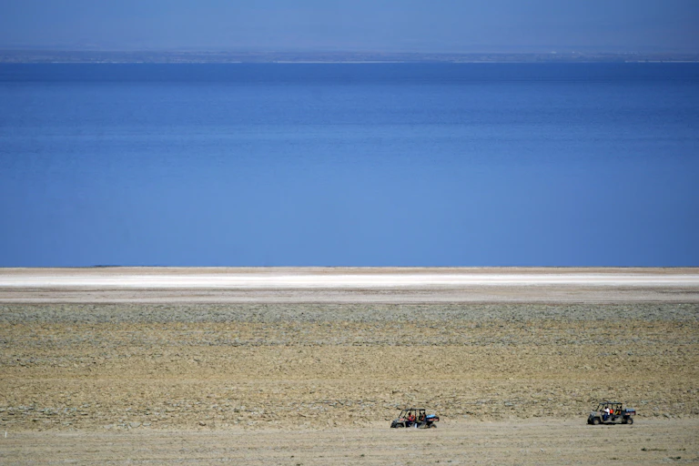 ATVs are driven along the shore of a blue body of water.