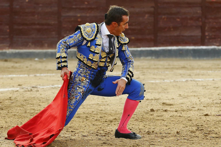 A matador kneels with a red flag during a bullfight.