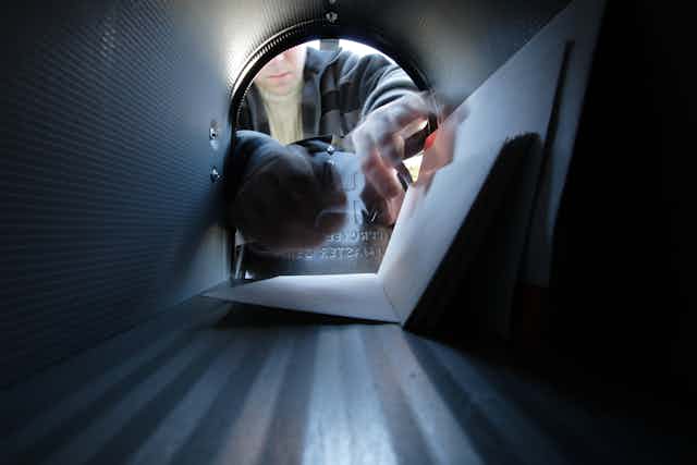 A person reaches into a mailbox, which contains several letters, as seen from perspective of inside mailbox