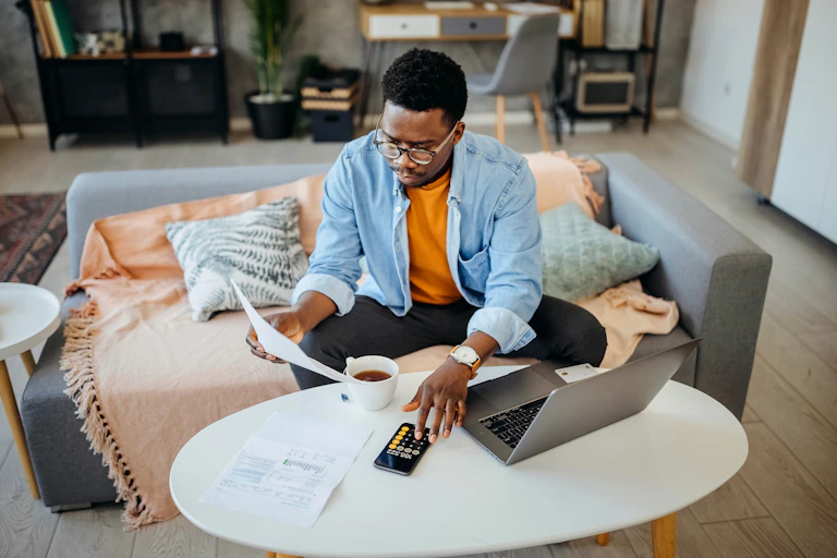 A Black man holds a paper while sitting on a couch. Next to him is a desk with a calculator, laptop and coffee mug.