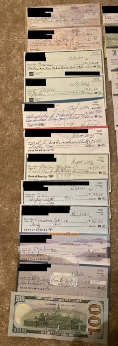 A dozen filled-in checks are displayed and slightly overlapping one another, with the back of a $100 bill at the bottom. The names and addresses are blacked out to protect victims' identities.