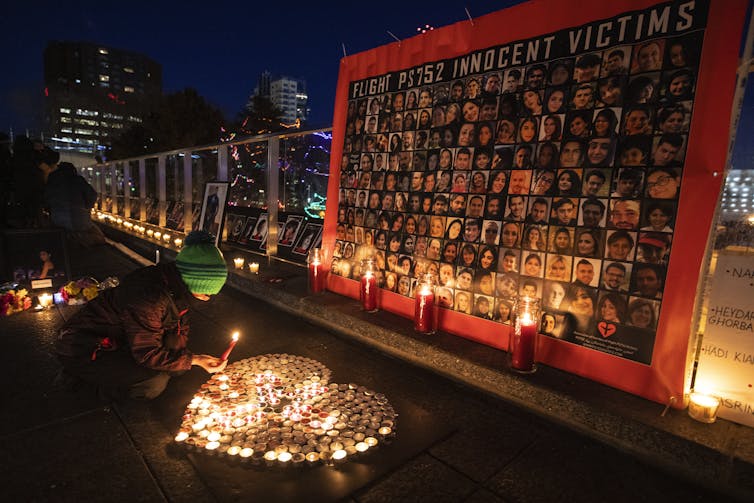 A person lights a candle in the dark outdoors in front of a photo montage memoria of victims of the Flight PS752 crash.