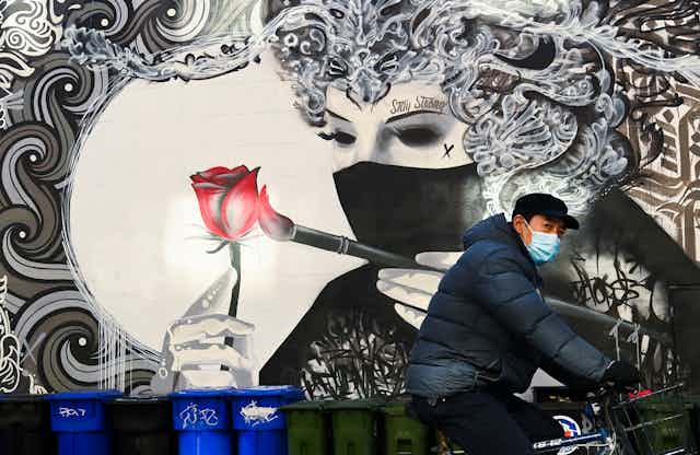 A man wearing a protective mask rides his bicycle past a street mural of a woman in a face mask painting a rose