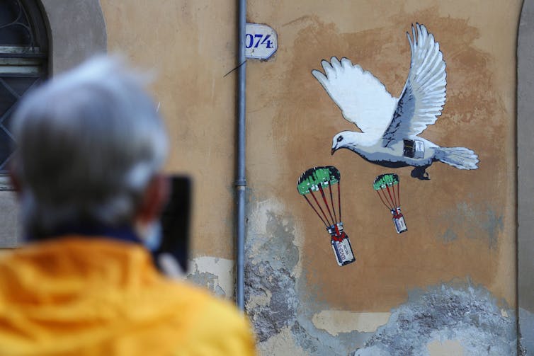Person seen from behind takes photo of mural of white dove dropping vials of COVID-19 vaccine