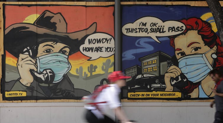 A cyclist walks past a painted mural depicting cartoon signs of people wearing face masks talking on the phone, checking out neighbors during the pandemic.