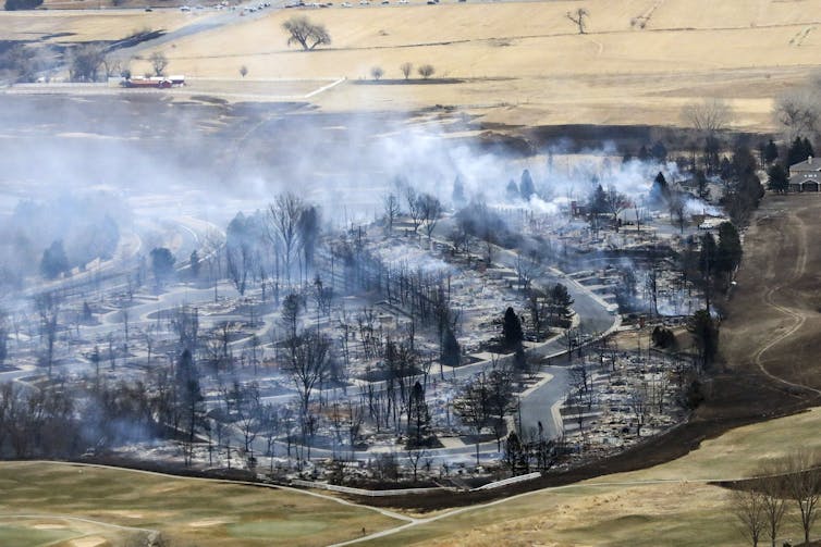 An aerial view of burned down houses and smoking trees, surrounded by pale yellow fields.