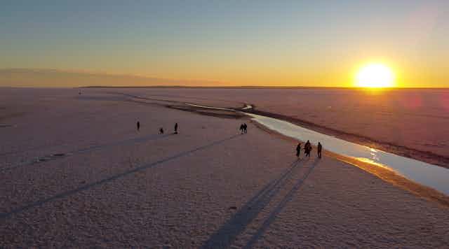 An aerial view of a thin sliver of water. People walk nearby as the sun hovers on the horizon.