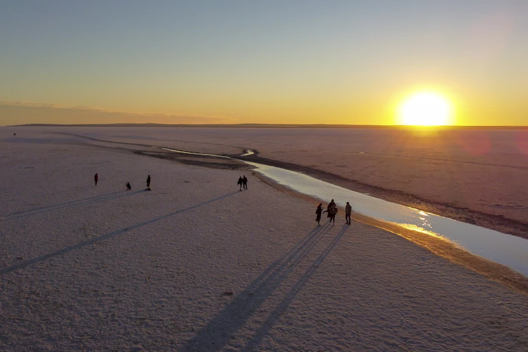 An aerial view of a thin sliver of water. People walk nearby as the sun hovers on the horizon.