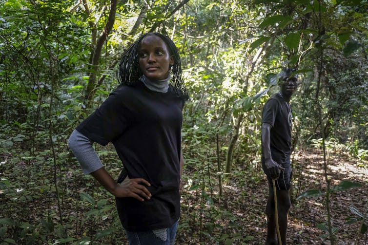 A woman stands in a forested area looking back, past the camera, over her shoulder. A man stands several metres in front of her, also looking back.