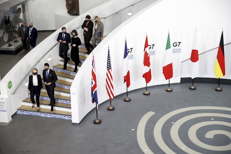 A group of people walk two by two down a winding staircase. At the bottom of the stair case are seven flags representing each of the G7 countries.